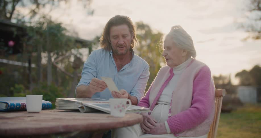 A middleaged man speaks with an elderly woman while showing her a photo from her photo album as they sit together at a table outdoors during the day