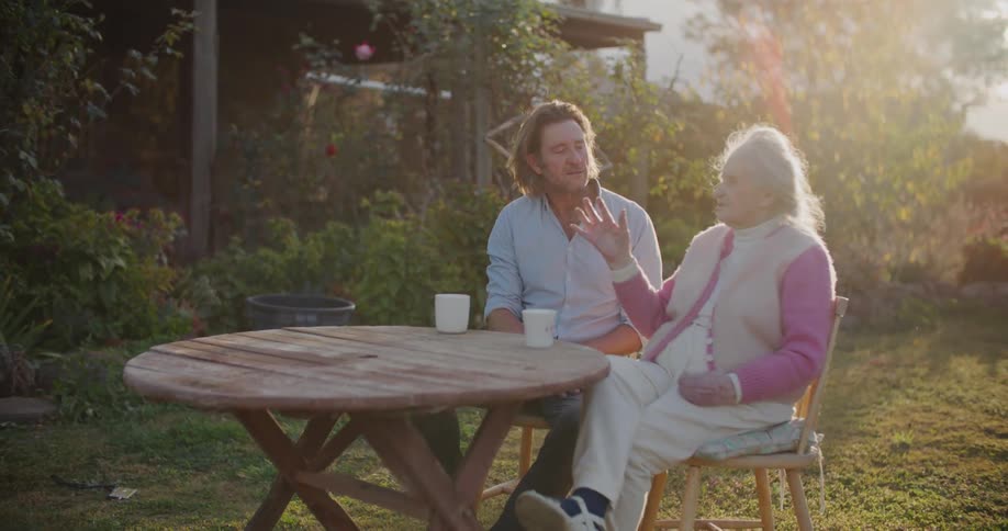 Middle aged man with a light blue shirt and elderly woman sit at table and talk to each other in the garden