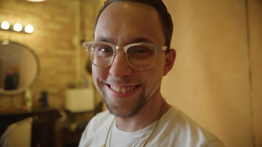 A man with glasses in a room with a wall and a mirror in the background