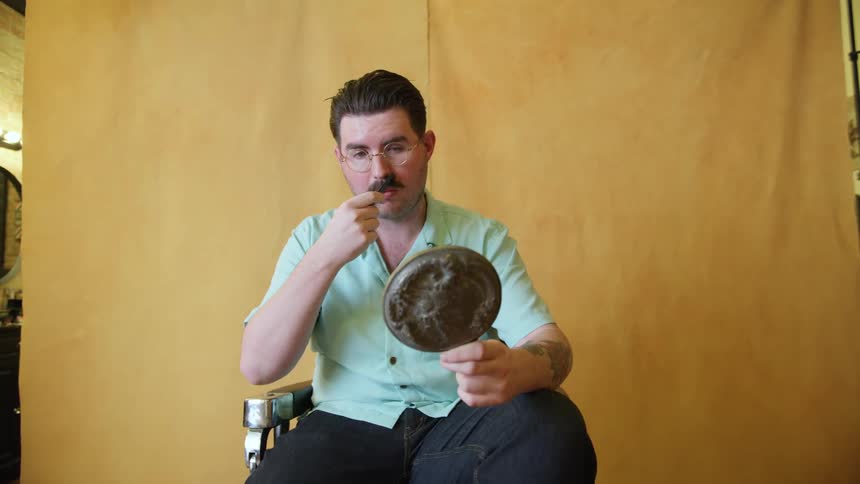 Man sitting on a chair inside a salon looking at the mirror combing his mustache