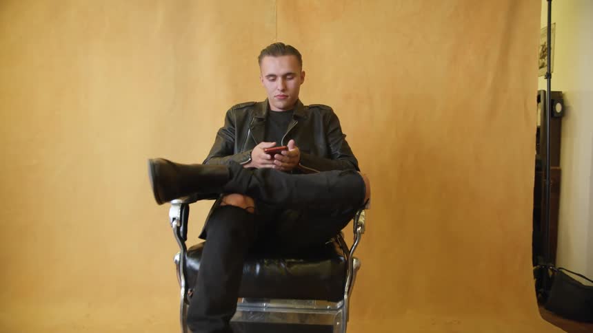 Man waiting in chair near cloth dividing him from beauty salon chairs takes out mobile phone
