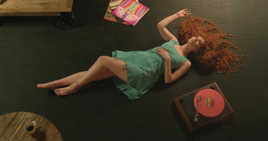Red headed woman lays on the floor twirling her hair while a record plays on a record player nearby