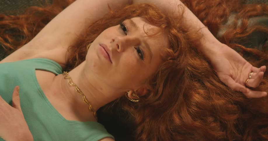 Woman with curly red hair lies down on the ground and moves her head