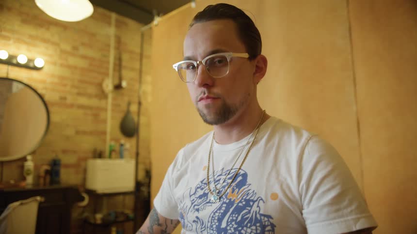 A man sitting in a barber chair gesture at the camera before looking over his shoulder