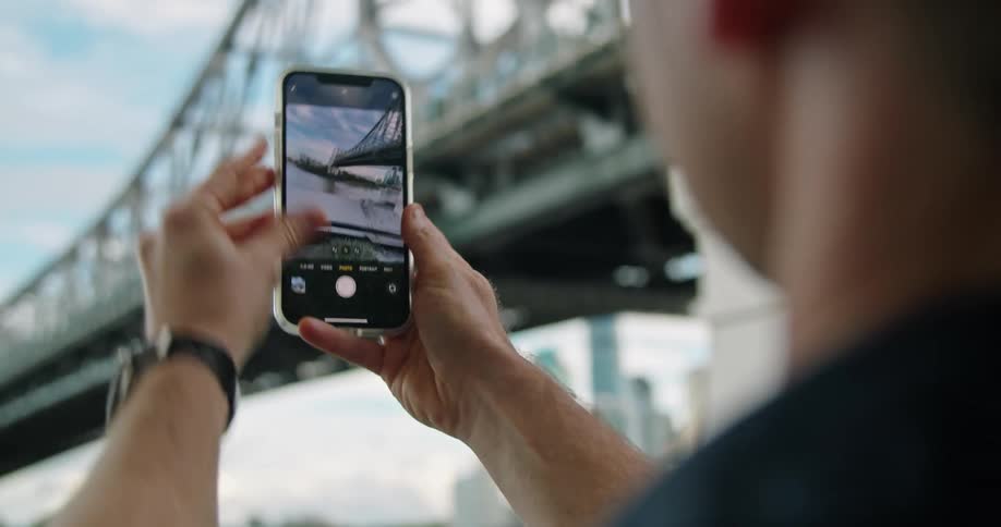 Man uses mobile phone to take pictures of bridge with city skyline in background