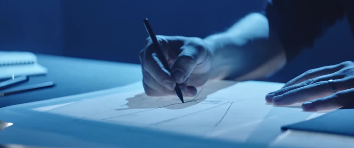 A man with glasses in a room drawing on a paper with a pen