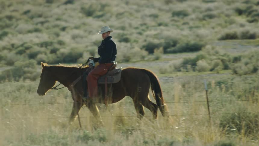 Lone cowboy wearing a cowboy hat, jean jacket and gloves riding a horse in front of a barb wire fence in Montana