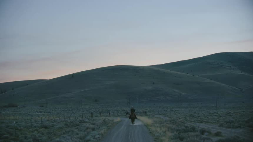 A man riding a horse on a dirt road with hills the sky and clouds in the background
