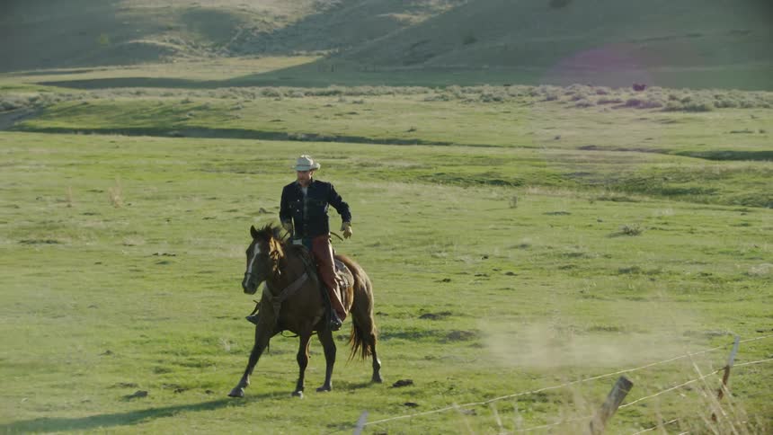A young cowboy wearing a cowboy hat and jean jacket riding a horse with hills in the background
