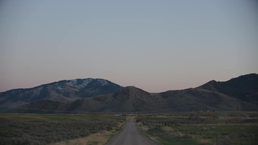 Pre sunrise light spills into a picturesque mountain valley as the pickup truck drives straight into the frame towards the ranch