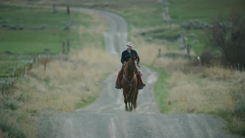 Static shot of a rugged young cowboy riding his horse on a rolling dirt road deep in the western United States