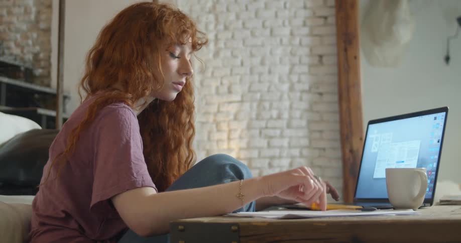 A teenager sits on the floor in her living room and takes notes while using her laptop
