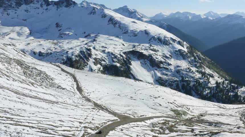 Black truck travels down a road alongside snow covered mountains and a mountain range on the horizon
