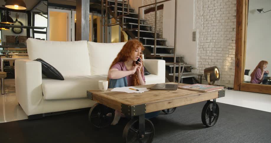 Young redhead woman sitting on the living room floor talking on her phone while using a laptop