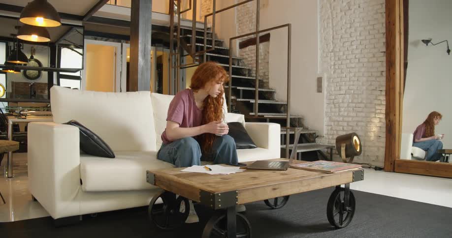 A teenage girl sits on a couch at home holding a cup of coffee and looks at her laptop