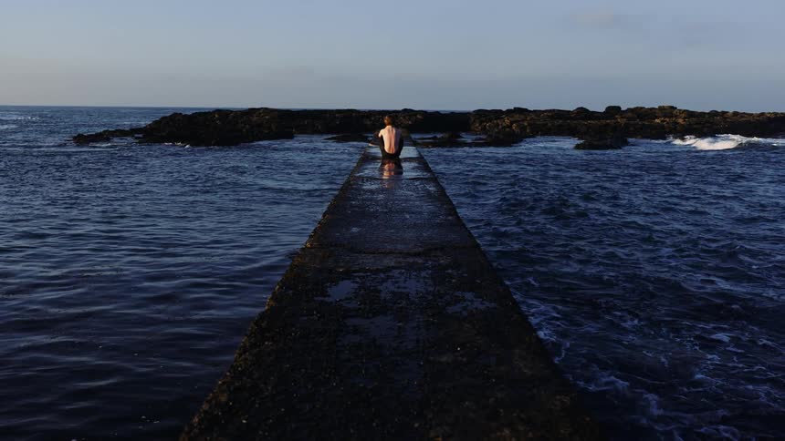 A man is sitting at the end of the pier in the water