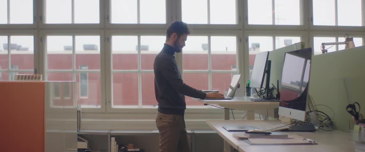 A man walks to an office computer and sets up his laptop in front of it