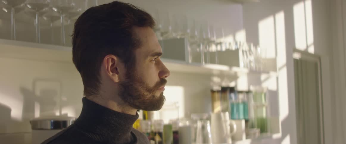 A man drinking from a mug while standing in front of a shelf of glassware