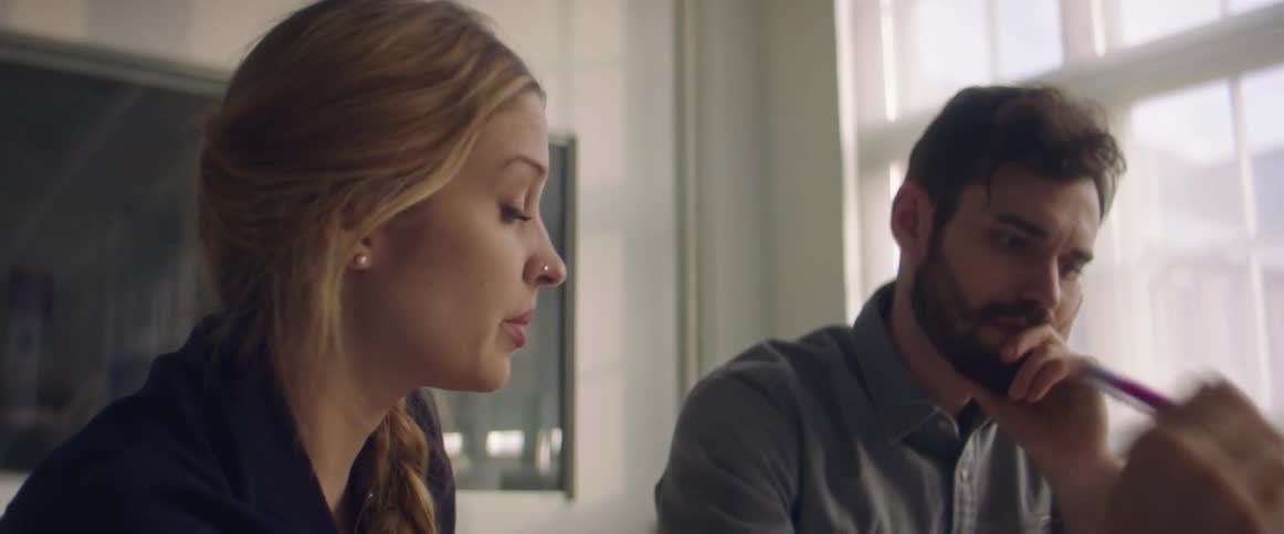 Blonde woman with a nose piercing sitting at table talks to man with a beard and a grey shirt in a room