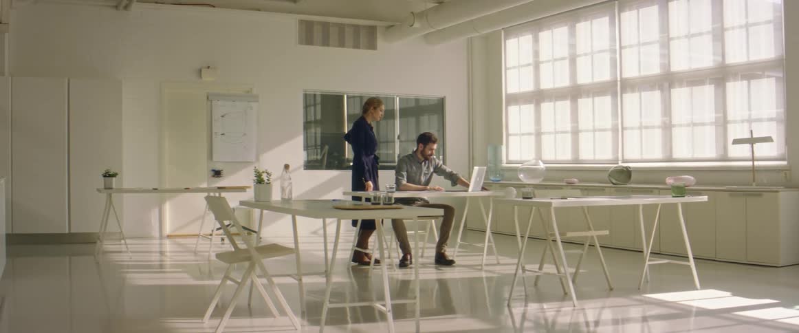 A man sits at a desk with a laptop and a woman stands behind him