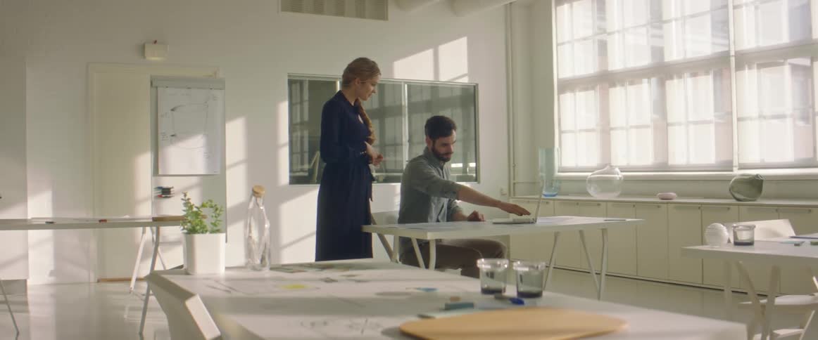 A man sits at a table with a laptop and talks to a woman standing behind him
