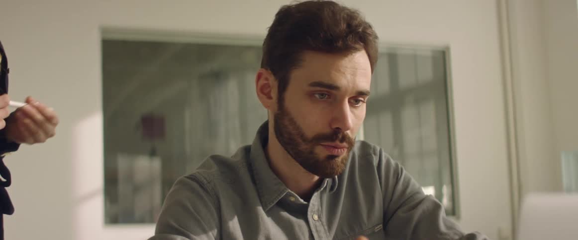 A man sitting before a laptop speaks to a woman standing next to him indoors during the day