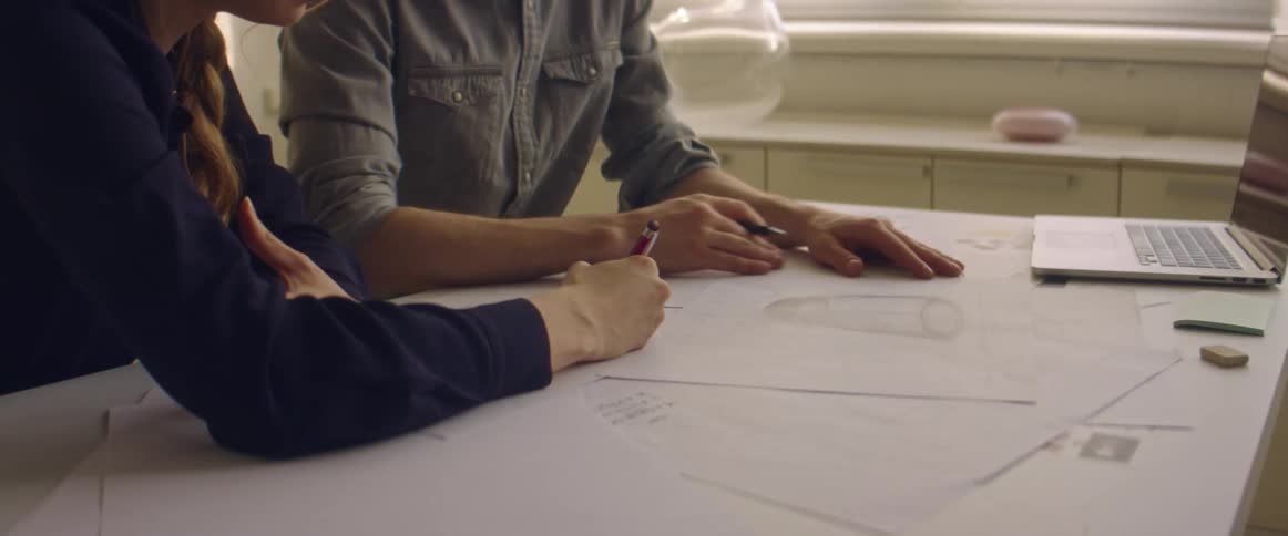 Man woman draws on a paper while sitting at a desk while a man sits next to her and a laptop sits on the desk