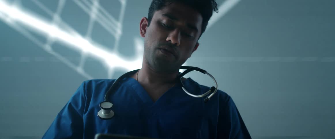 A physician looks down at his tablet computer while abstract images flash on a large screen behind him