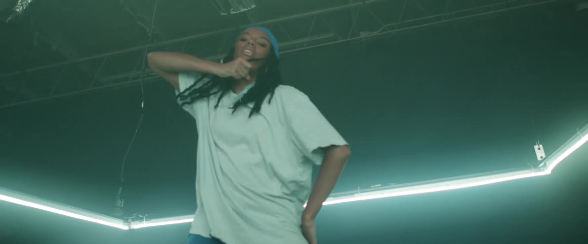 Woman with a blue headband braids hoop earrings and a white loose t shirt dances in a dark room with lights