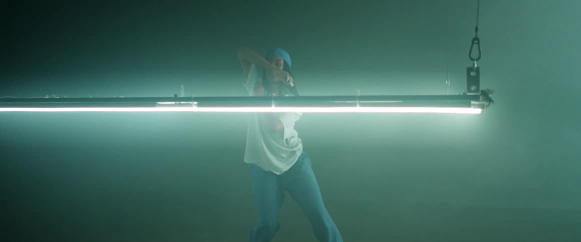 Woman with a blue headband braids hoop earrings and a white loose t shirt dances in a dark room with lights