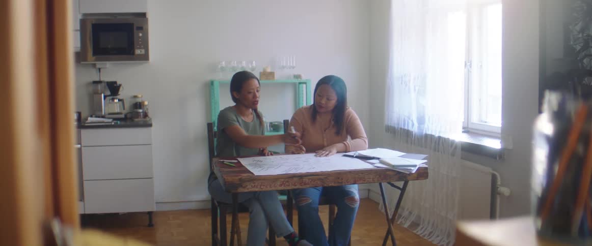 Two women sit together in a kitchen while discussing and reviewing plans together