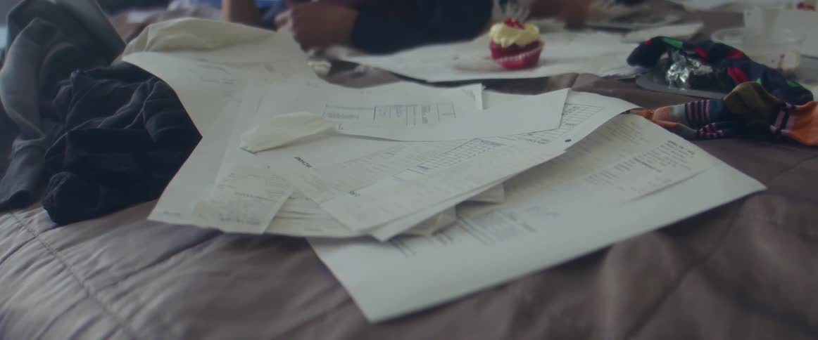 Woman lies on bed next to cupcake and strewn paperwork while watching laptop