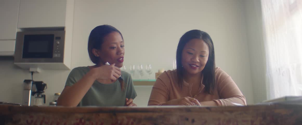 Two woman sit at a table with paper and pens working on something together