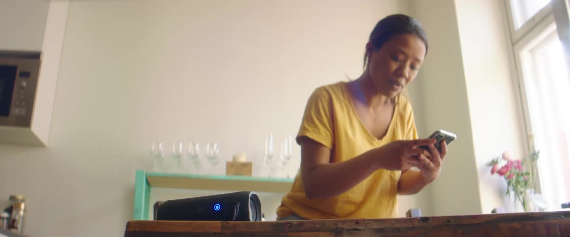 A woman leaning over a table shifts her attention to her smart phone while looking at its screen