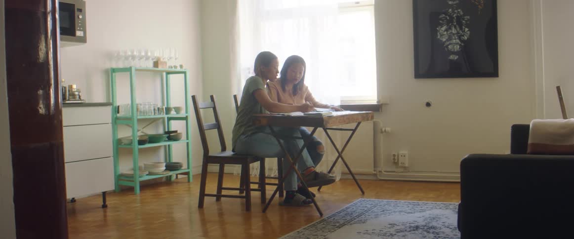 Two women in a room sitting on chairs at a table with a wall and window in the background