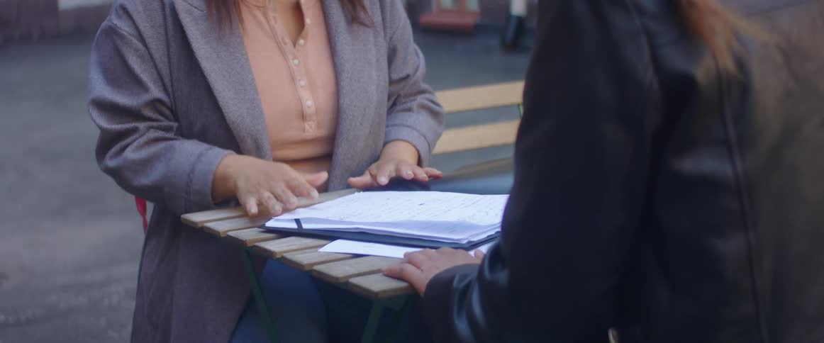 Two women sitting on chairs at a table with a building with doors in the background