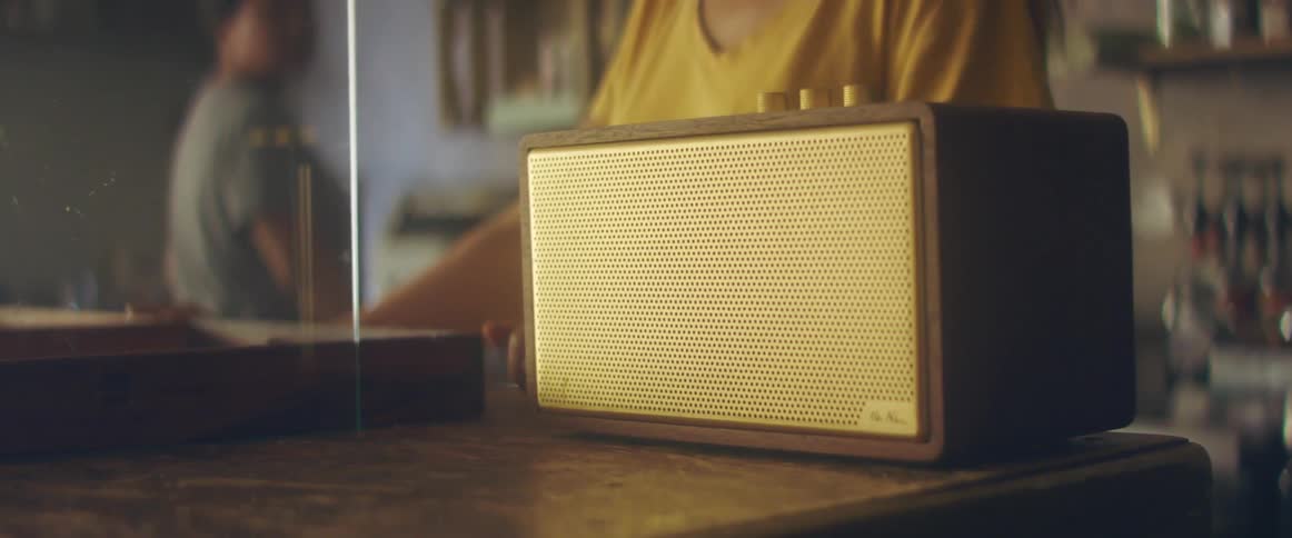 Two women in a room with one turning a knob on a radio with a wall and shelves in the background