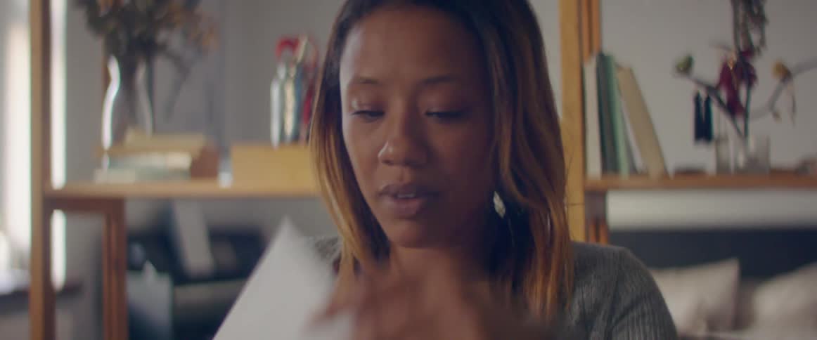 A woman carrying a box takes a seat in a bedroom and looks at a sheet of paper