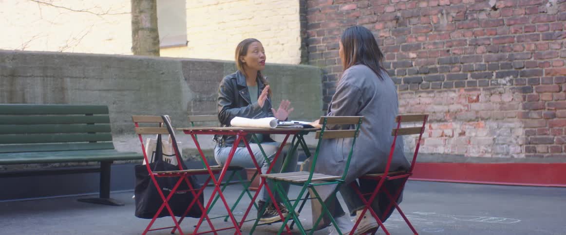 Two women sitting at a small table outdoors have a lively conversation
