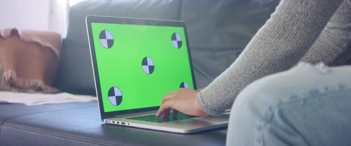 A person in a room sitting on a couch and typing on a laptop with a wall and window in the background