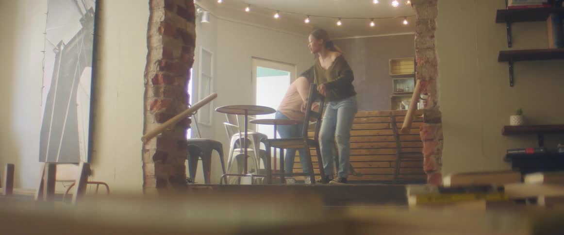A pair of women arrange furniture indoors during the day