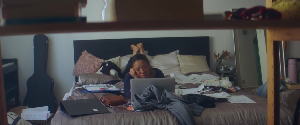 A woman in a bedroom laying in bed looking at a laptop with a wall in the background