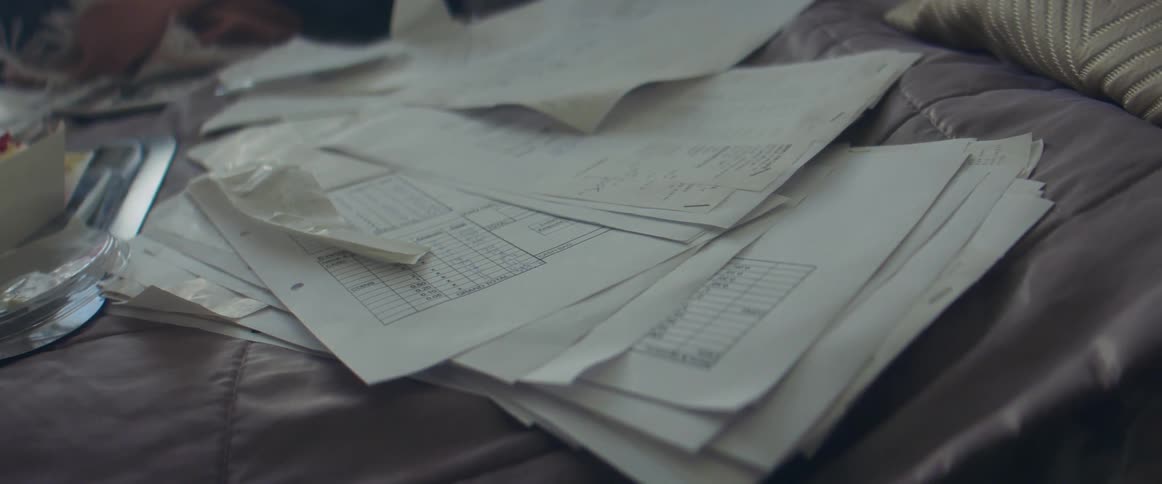 A pile of papers is left atop a bed next to a takeout box with food in it