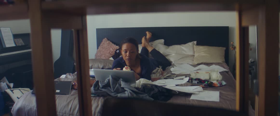Woman with a ponytail and in a purple pyjama lies on bed eats dessert and looks at laptop in bedroom