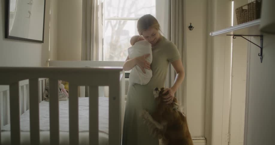 A woman pets a dog standing against her leg while holding a baby indoors during the day