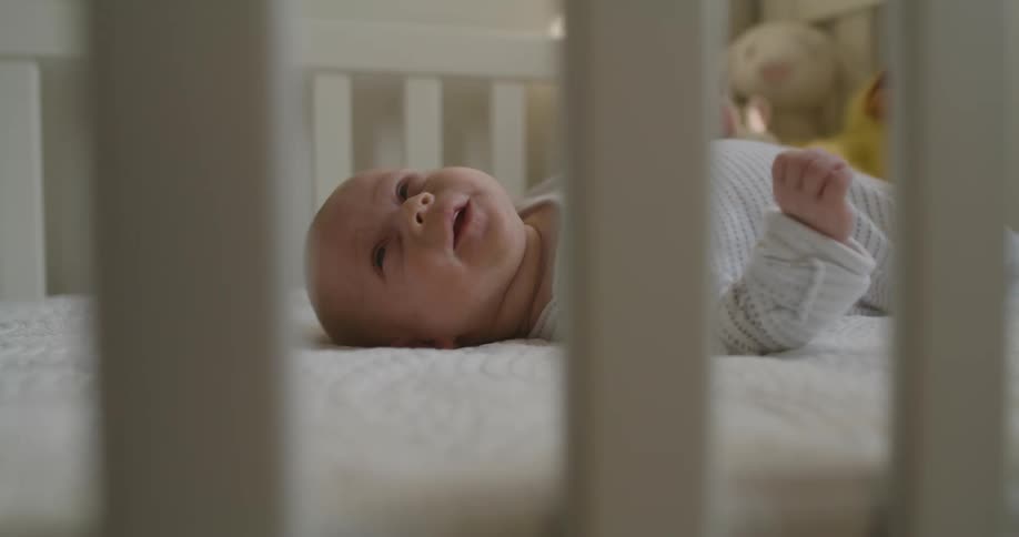 A baby in a bedroom laying on a crib with a wall and toys in the background
