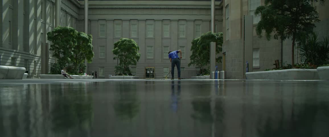 A man with a broom sweeping the floor with a building and trees in the background