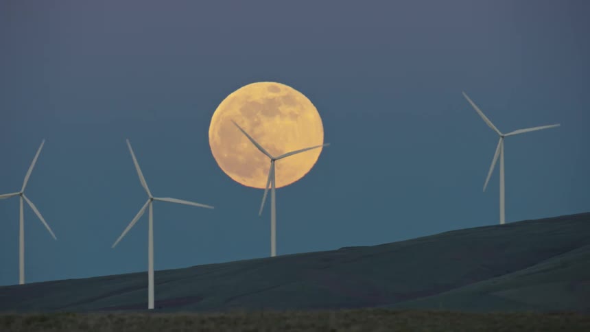 Dolly shot of a super moon rising above four whirling green energy renewable wind turbine electricity generators