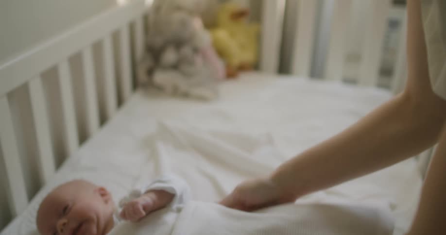 A woman in a bedroom wrapping a baby in a blanket in a crib with a wall and window in the background