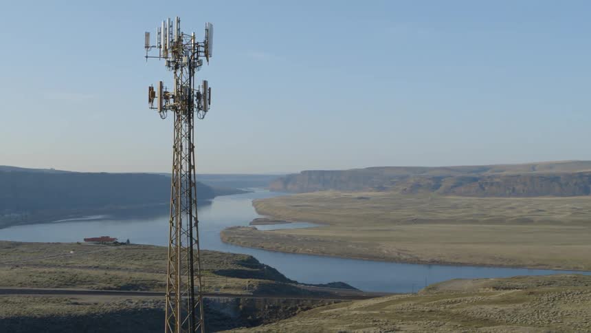 Vehicles travel on a rural highway near a winding river as a cell phone tower works overhead